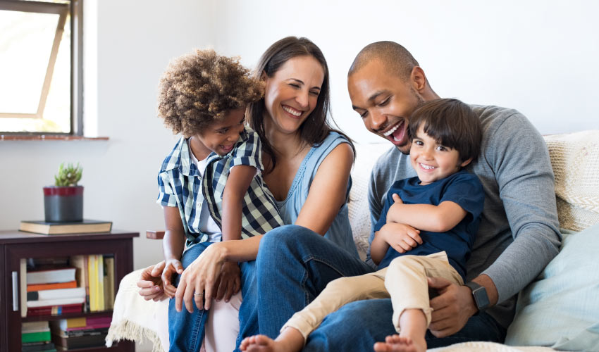 Multiracial family of mom, dad, and two sons smile and laugh while sitting on a couch together
