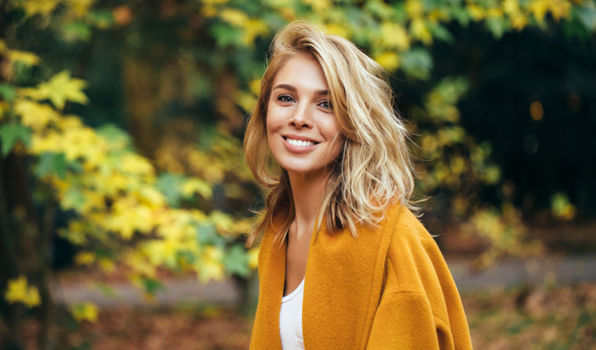 Blonde woman with dental implants smiles while wearing a golden pea coat outside by the fall leaves