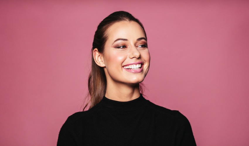 Brunette woman in a black shirt smiles against a pink background after overcoming her dental anxiety