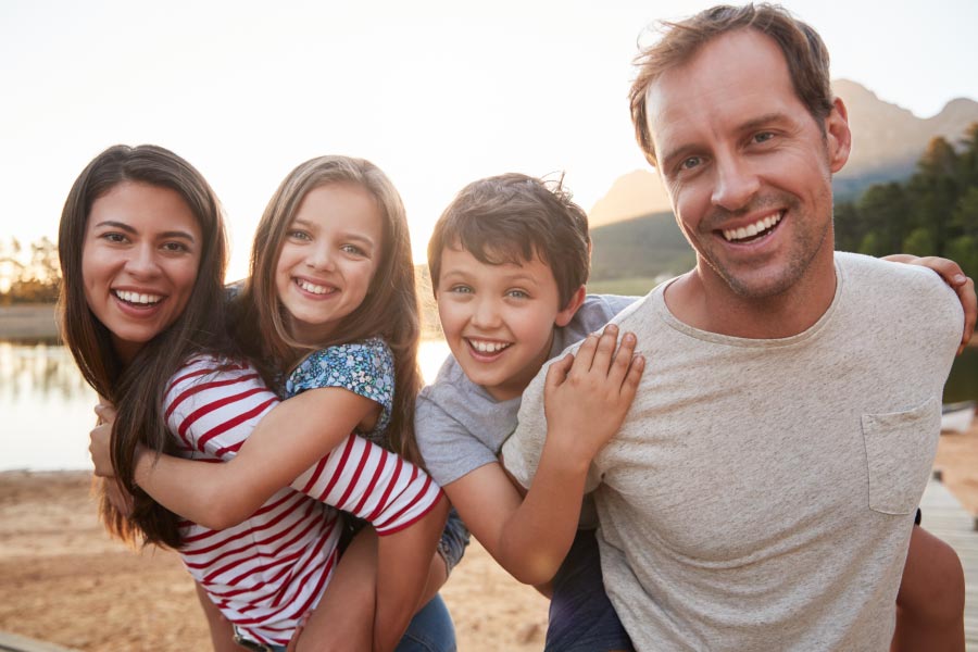 Young family standing in front of a lake with a girl on the mother's back and a boy on the father's.