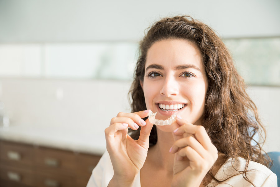 Curly haired smiling woman holding an Invisalign clear aligner in front of her chin.