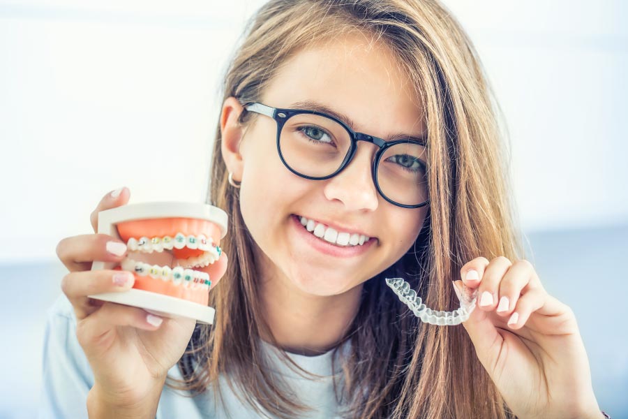 Young teen holding an Invisalign clear aligner in one hand and a mouth model with braces in the other.